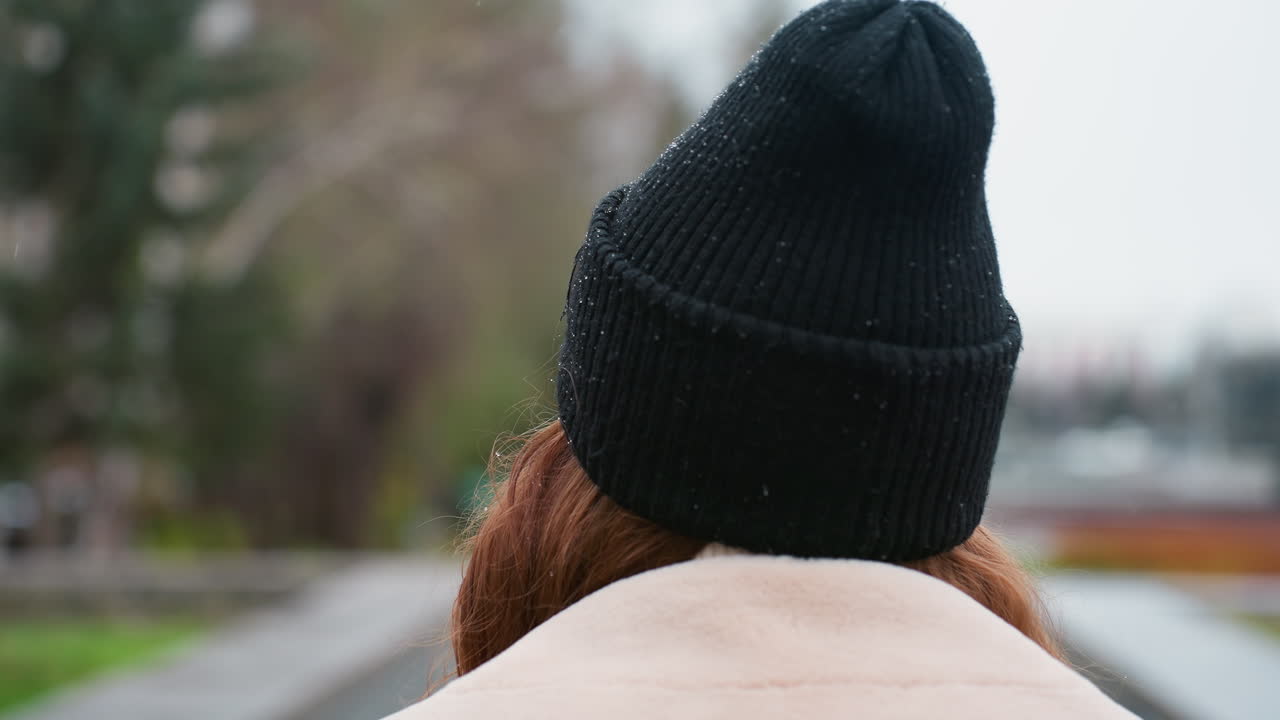 Close back view of lady walking outdoors in black knit cap and beige collar winter jacket, blurred park background and overcast sky create peaceful mood as she strolls away from camera quietly