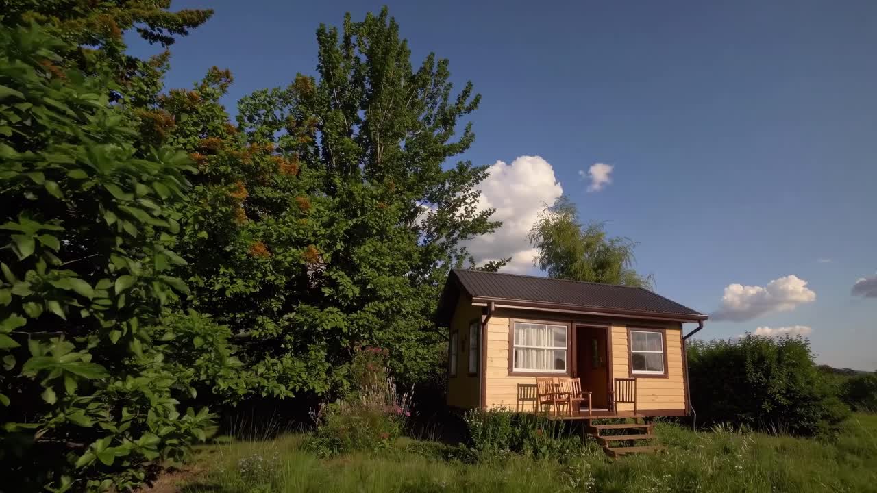 Wide-angle video shot of a small wooden cabin in a lush green landscape, with a clear blue sky