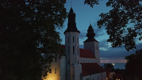 sensational tree framed picturesque scene of visby cathedral at dusk with glowing lights on church and boat in ocean sea water in background, sweden, handheld