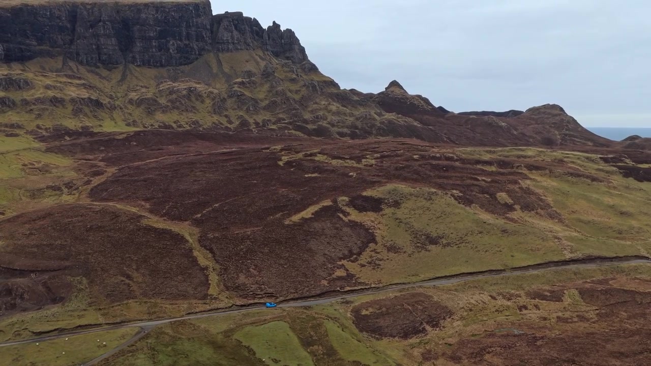 Vista aerea di un'auto blu che percorre una strada tortuosa attraverso il suggestivo paesaggio del Quiraing sull'Isola di Skye in Scozia.