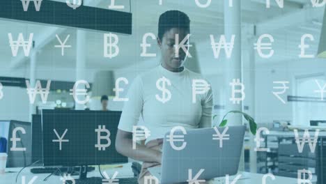 Animation-of-currency-symbols-over-african-american-woman-sitting-on-desk-and-using-laptop-in-office