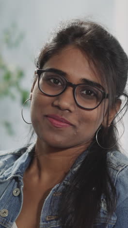 young indian woman puts on glasses with black rim looking in camera and smiling. portrait of female person with ponytail on blurred background closeup