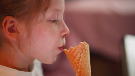 close up of young girl eagerly eating ice cream cone indoors, smiling with creamy mouth, wearing white shirt, soft pink blurred background visible behind