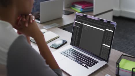 Mixed-race-woman-sitting-at-desk-watching-coding-data-processing-on-laptop-screen