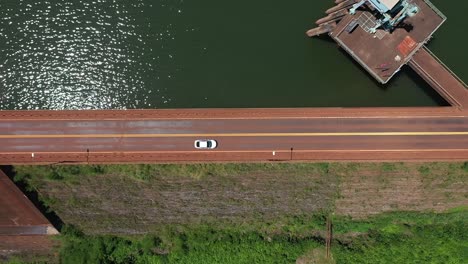 Vista-Aérea-De-Drones-De-Un-Solo-Vehículo-Cruzando-Una-Presa-De-Un-Embalse-A-Través-Del-Cual-Pasa-Una-Carretera-Tropical-En-Misiones,-Argentina