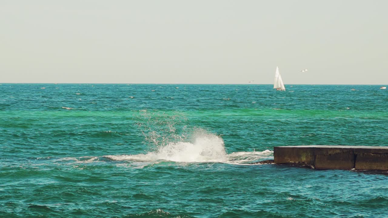 A sailboat, yacht on the horizon in the sea. Splashing waves, ship in the background. Breakwater.