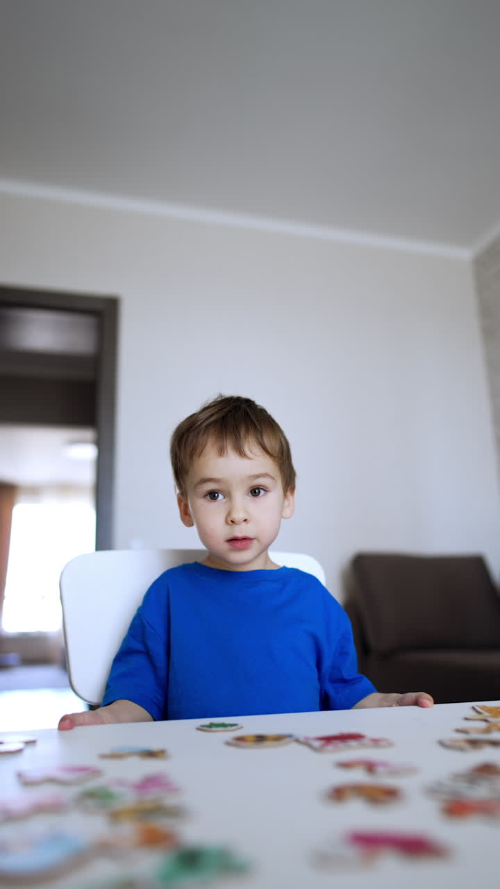 Beautiful Caucasian baby boy sitting at desk playing a game. Cheerful kid collects puzzles. Vertical video.