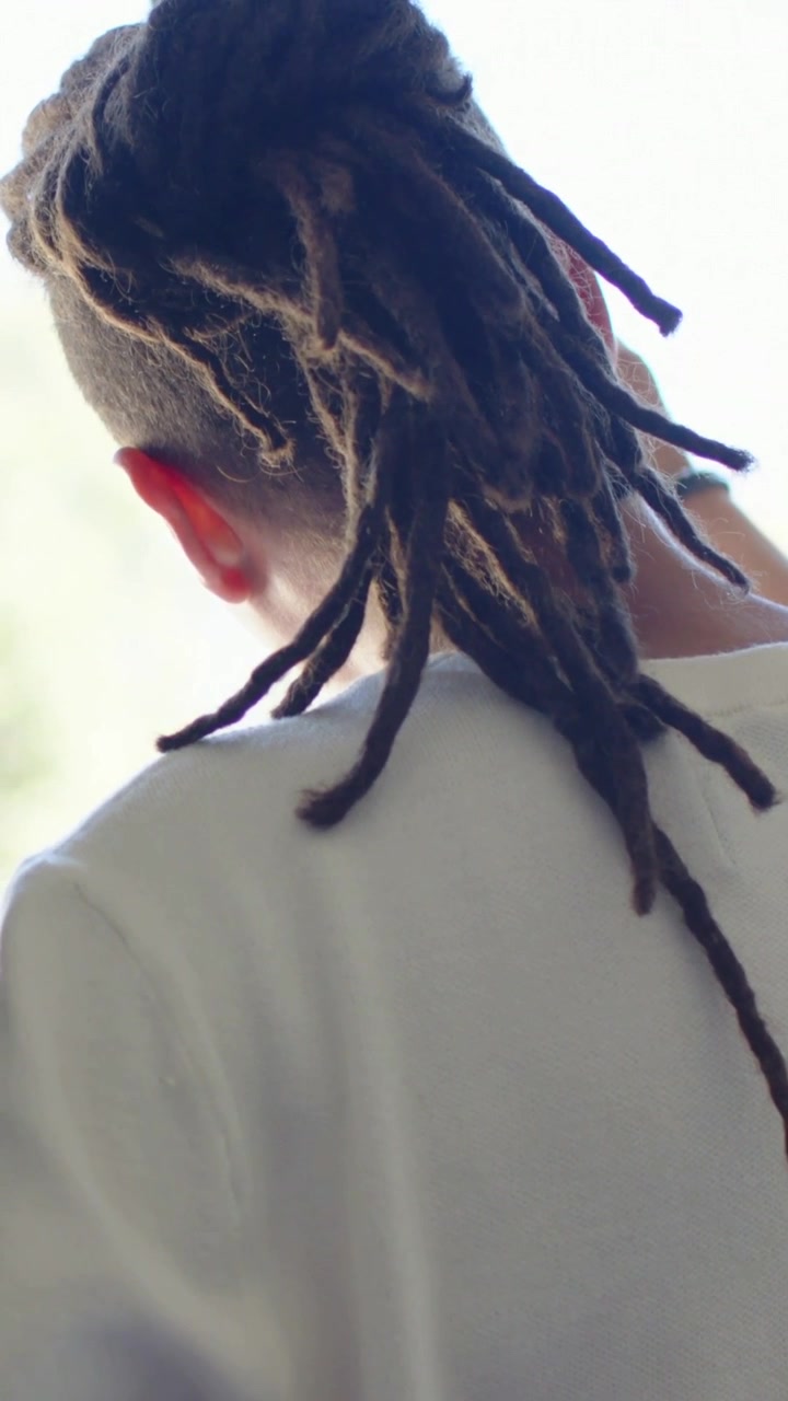 Vídeo vertical: Protegendo-se do sol forte, homem com dreads ajeitando o cabelo na janela, camiseta branca.