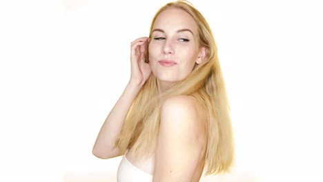 against a plain white studio background, a young woman with blonde hair looks over her shoulder and smiles