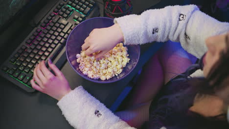 overhead view of programmer in fluffy sleeve using colorful keyboard while working, bowl of popcorn placed beside red condenser microphone on dark desk in studio with relaxed ambient lighting