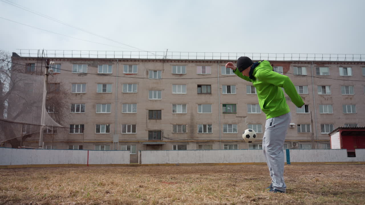 갈색 필드에서 네온 재킷을 입은 백인 남성이 공을 저글링하고 컨트롤하는 단독 플레이어 훈련, 배경에는 도시 아파트 단지, 집중적인 반복 및 타이밍 훈련, 흐린 조명