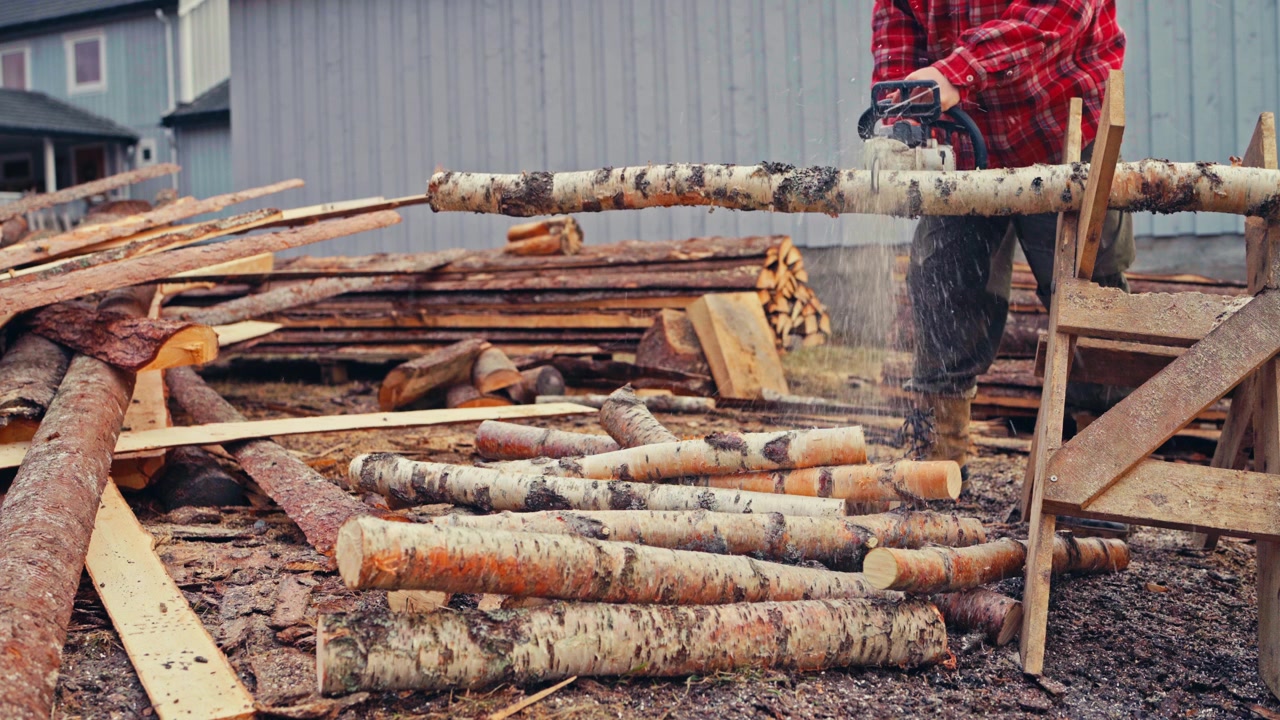 Man Uses A Chainsaw To Cut A Birch Log Resting On A Wooden Sawbuck Beside Scattered Timber On A Rural Farmstead In Norway. - wide shot
