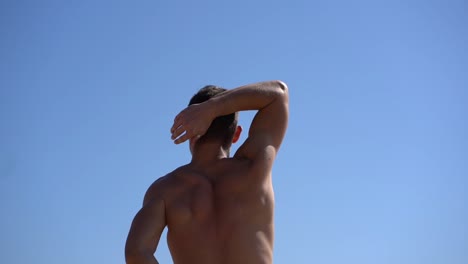 muscular shirtless man stretching arms against blue sky