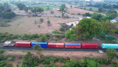 aerial rotating drone view of freight train with red, blue coloured carriages running along a highway with green fields and trees and urban vegetation near vadodara city in india