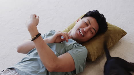 asian man relaxing on floor with dog, smiling and enjoying playful moment, at home