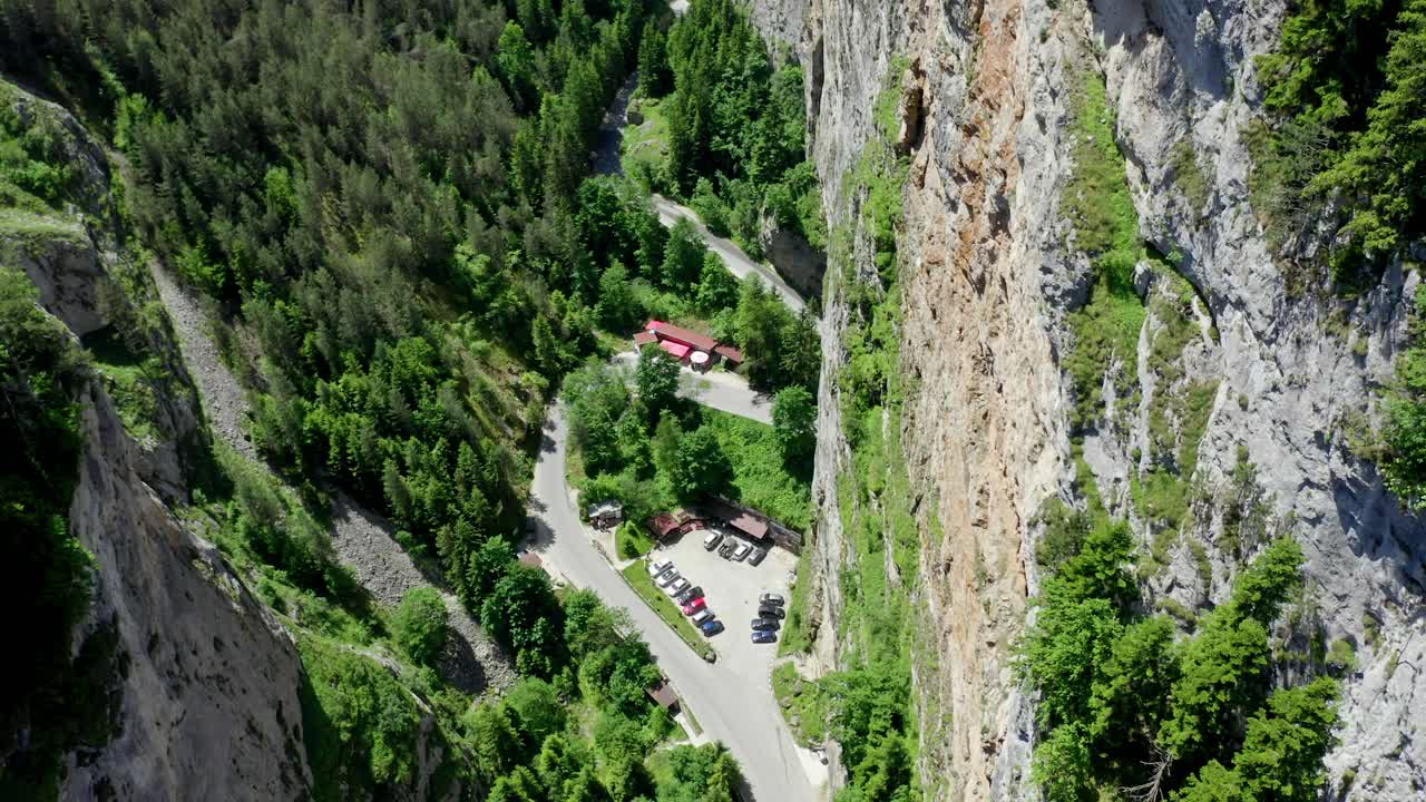 el dron desciende revelando una cara de roca dentada en la parte derecha del cañón y el espacio de estacionamiento debajo con edificios para los visitantes que vienen, trigrad gorge, provincia de smolyan, bulgaria