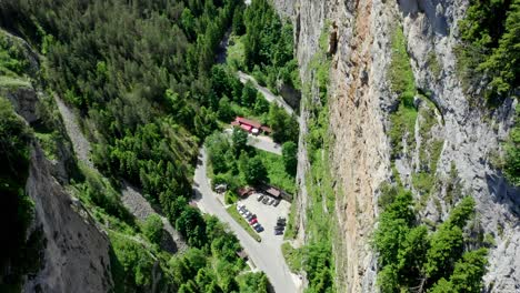 El-Dron-Desciende-Revelando-Una-Pared-Rocosa-Irregular-En-La-Parte-Derecha-Del-Cañón-Y-El-Espacio-De-Estacionamiento-Debajo-Con-Edificios-Para-Los-Visitantes,-Garganta-De-Trigrad,-Provincia-De-Smolyan,-Bulgaria