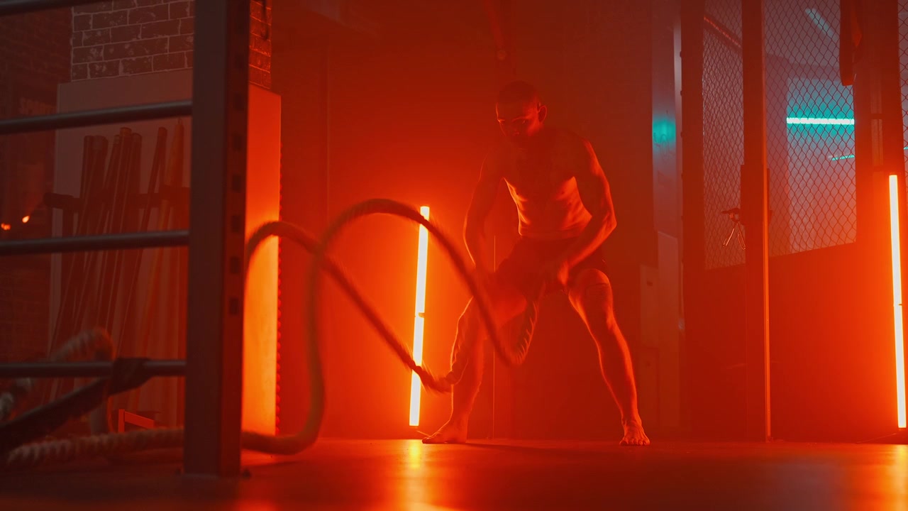 Muskulös man utför intensiv battle rope-träning i ett rött upplyst gym