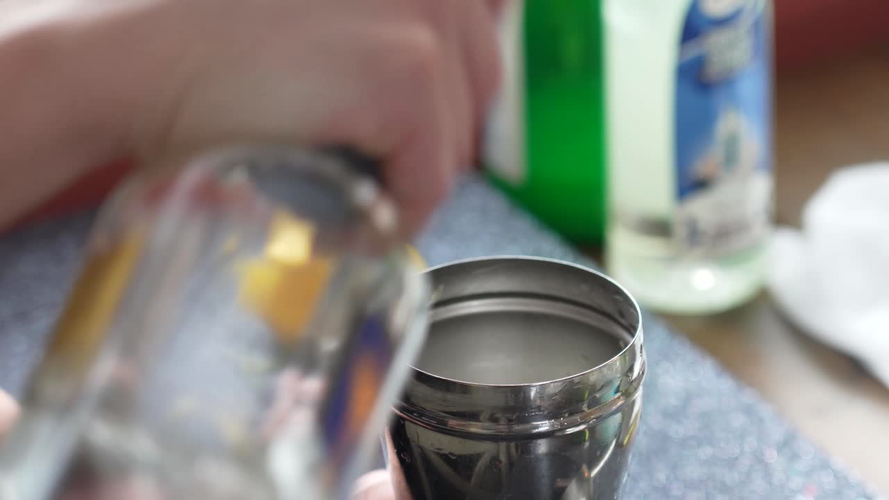 Bartender Pours Clear Liquid Alcohol Vodka, Rum, Tequila, Gin or Simple Syrup into Cobbler Shaker to make a Cocktail or Mixed Drink. Closeup of Woman’s Hands with a Lemon and Bottles in Background