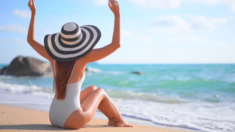 Una-Mujer-Joven-En-Traje-De-Baño-Y-Un-Sombrero-Flexible-Para-El-Sol-Se-Sienta-En-Una-Playa-De-Arena-Levantando-Los-Brazos-Para-Dar-La-Bienvenida-Al-Surf-Entrante