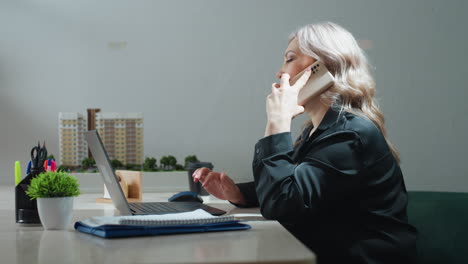 side view of professional woman sitting at desk in modern office picking up phone call while multitasking on laptop, surrounded by coffee cup, desktop supplies, property models, and documents