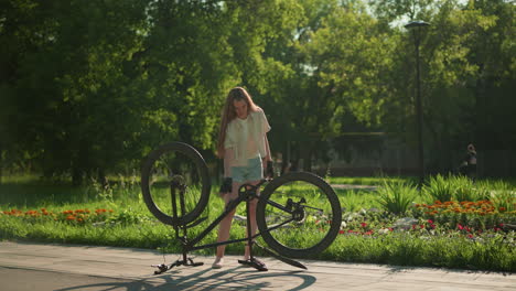 young woman rotates the pedal of an upside-down bike near a vibrant garden with blooming flowers, blurred background shows greenery and someone walking nearby