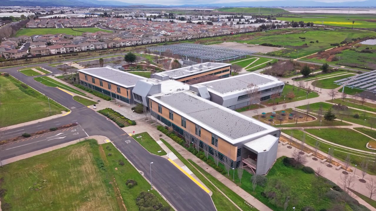 Riprese aeree con drone dall'alto inquadrano l'Ohlone College Newark Center sullo sfondo del più ampio paesaggio di Newark, enfatizzando l'accessibilità e la connettività urbana.