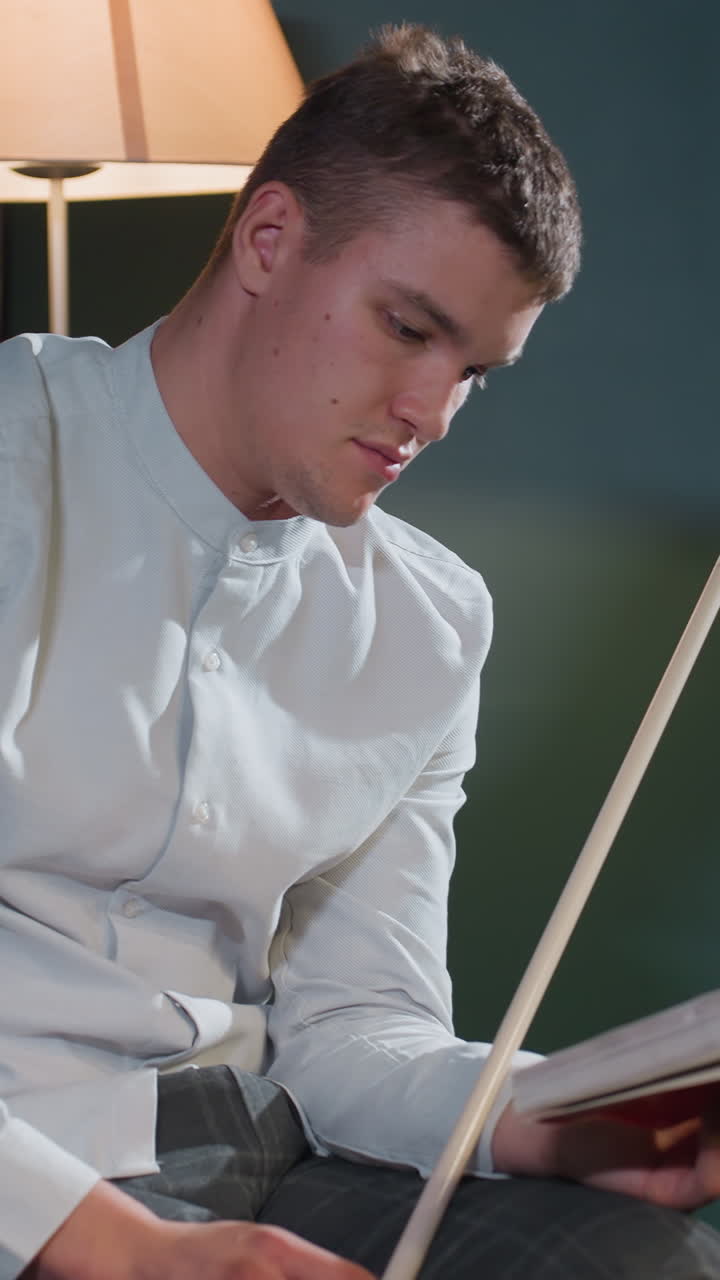 primer plano de un tipo con camisa blanca sentado y leyendo un libro mientras sostiene un palo con expresión enfocada. la iluminación cálida mejora la atmósfera intelectual y estratégica en un entorno interior moderno