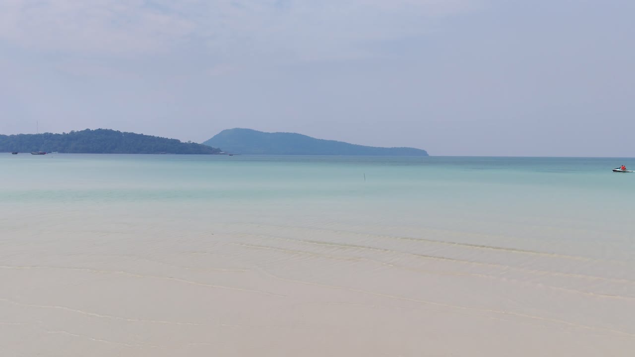 Tranquil shallow waters meet soft white sand under a hazy blue sky near Kaoh Rong Sanloem, Cambodia, with distant forested islands adding depth to the calm tropical seascape