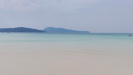 tranquil shallow waters meet soft white sand under a hazy blue sky near kaoh rong sanloem, cambodia, with distant forested islands adding depth to the calm tropical seascape