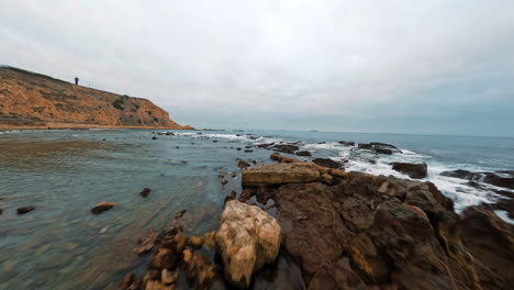 Vuelo-Bajo-Sobre-La-Superficie-Del-Agua,-Costa-Del-Mar-Con-Rocas-Que-Se-Elevan-Desde-El-Agua-Y-Escarpa-Empinada-A-Lo-Largo