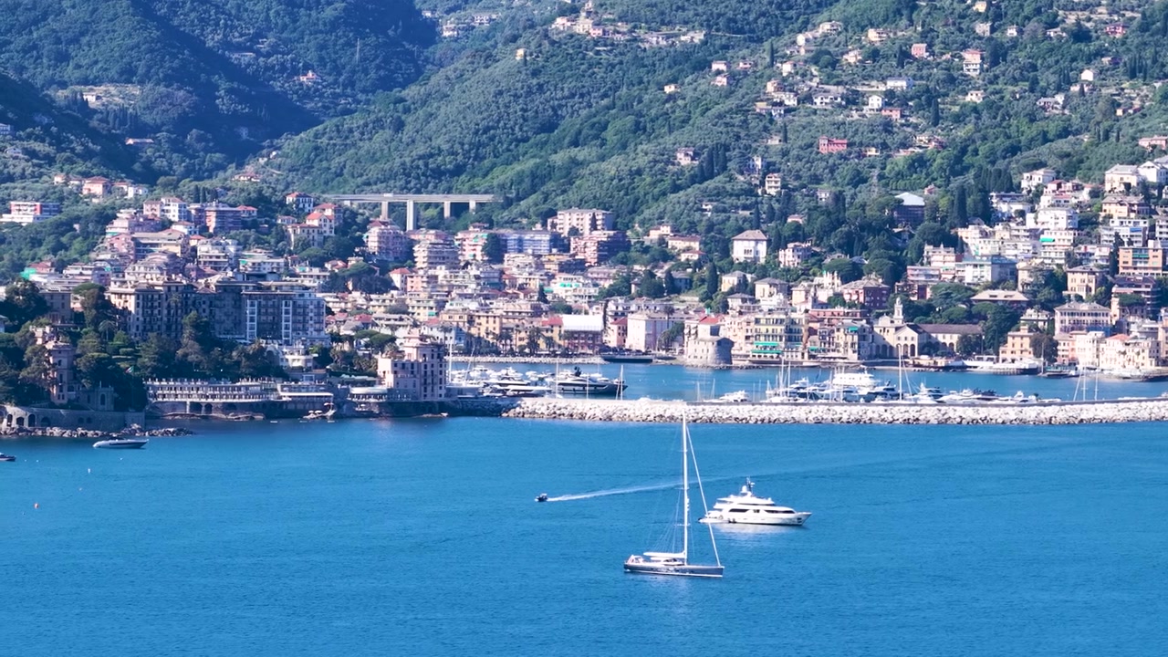 Barche a vela scivolano sull'acqua blu vicino a una storica città costiera italiana sotto un sole splendente.