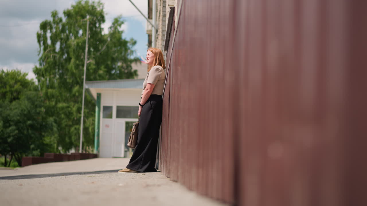 Side view of student holding bag walking toward building wall on sunny day, preparing to lean and rest, surrounded by green trees and brick architecture, symbolizing fatigue