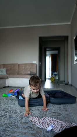 happy toddler boy gets into the big suitcase. kid stands up and picks some of his clothes scattered around the room. vertical video.