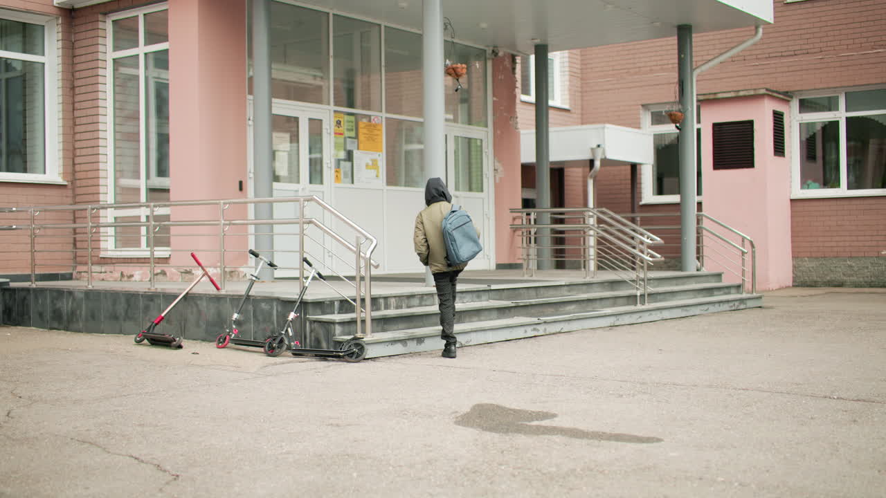 흐린 날, 학교 입구를 향해 걸어가는 뒷모습의 배낭을 멘 학생, 계단 옆에 주차된 스쿠터, 도시 캠퍼스 외부, 조용한 아침 등굣길, 교육 테마, 안전한 환경