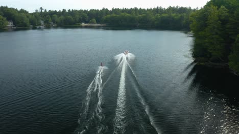 at midday, a woman in skis being towed by a motorboat is seen at a blue lake with splashes of water or waves, several lush, green trees in the background, a few dwellings