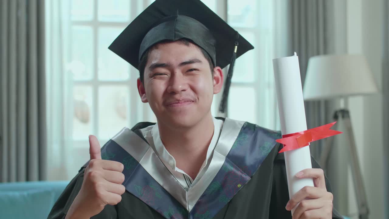 Excited Asian Man Holding A University Certificate, Smiling And Thumb Up To Camera. Male Graduate Wearing A Graduation Gown And Cap Sitting On The Living Room