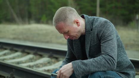 a close view of a man in a gray jacket and blue jeans, handling a handgun while sitting down and inspecting it beside a railway track, with a blur view of forest in the surrounding