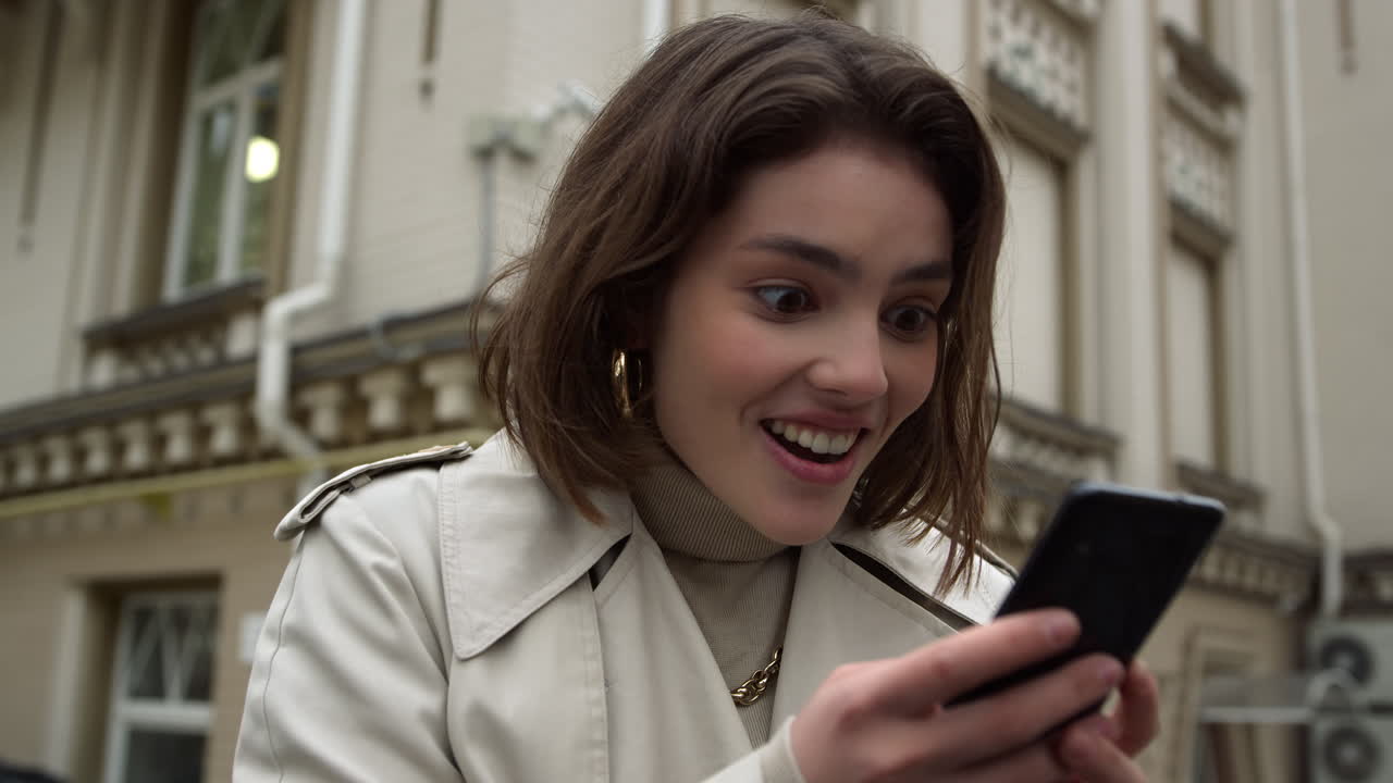 Excited woman reading message on phone outdoor. Girl holding cellphone in city.