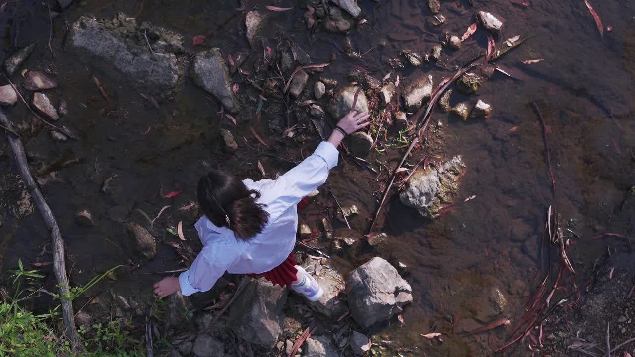 Pemandangan udara menangkap seorang gadis menyeimbangkan diri dengan hati-hati melintasi bebatuan di aliran sungai Warrandyte Park. Matahari senja menyoroti latar alami yang tenang dan gerakan dinamis.