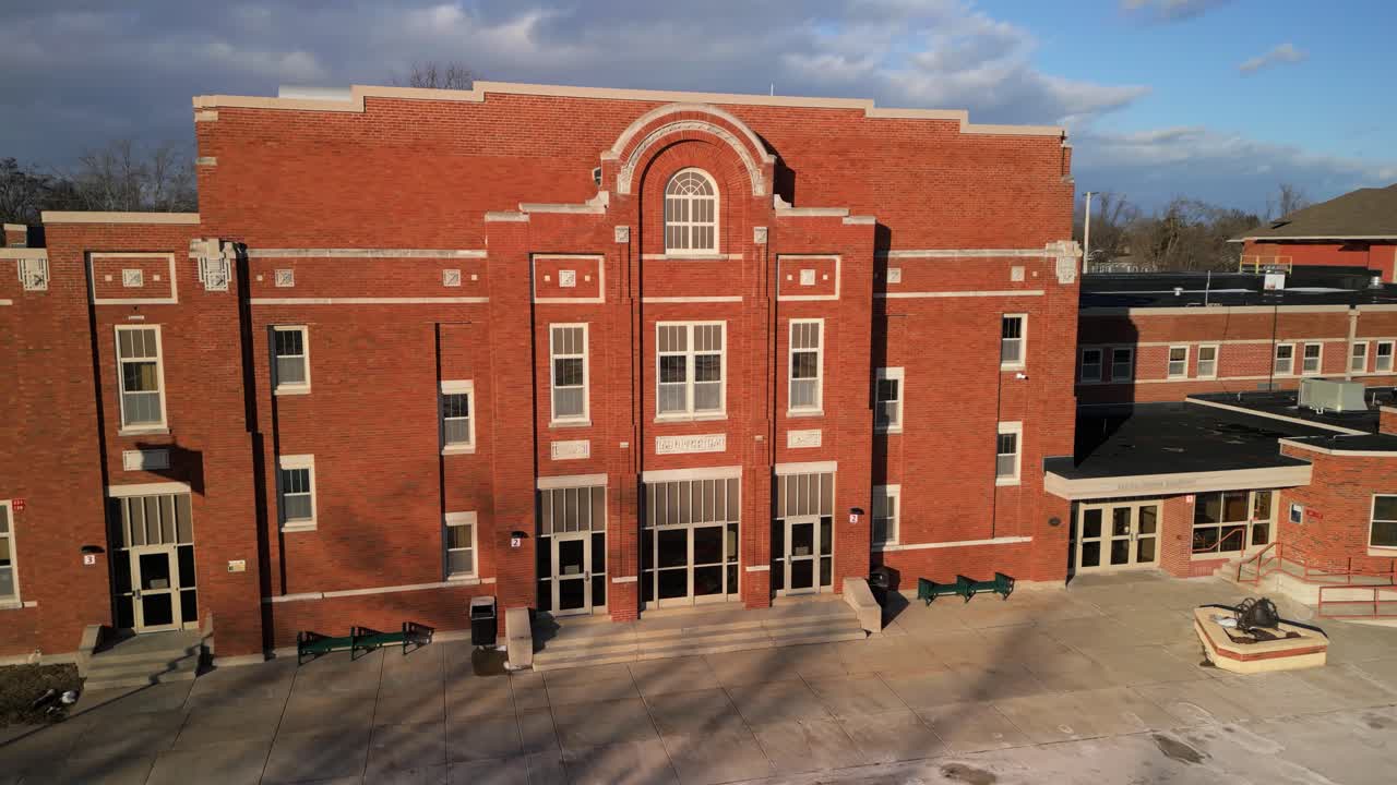 Front view of old suburban school with red brick facade, large entry, and daylight shadows, aerial angled approach