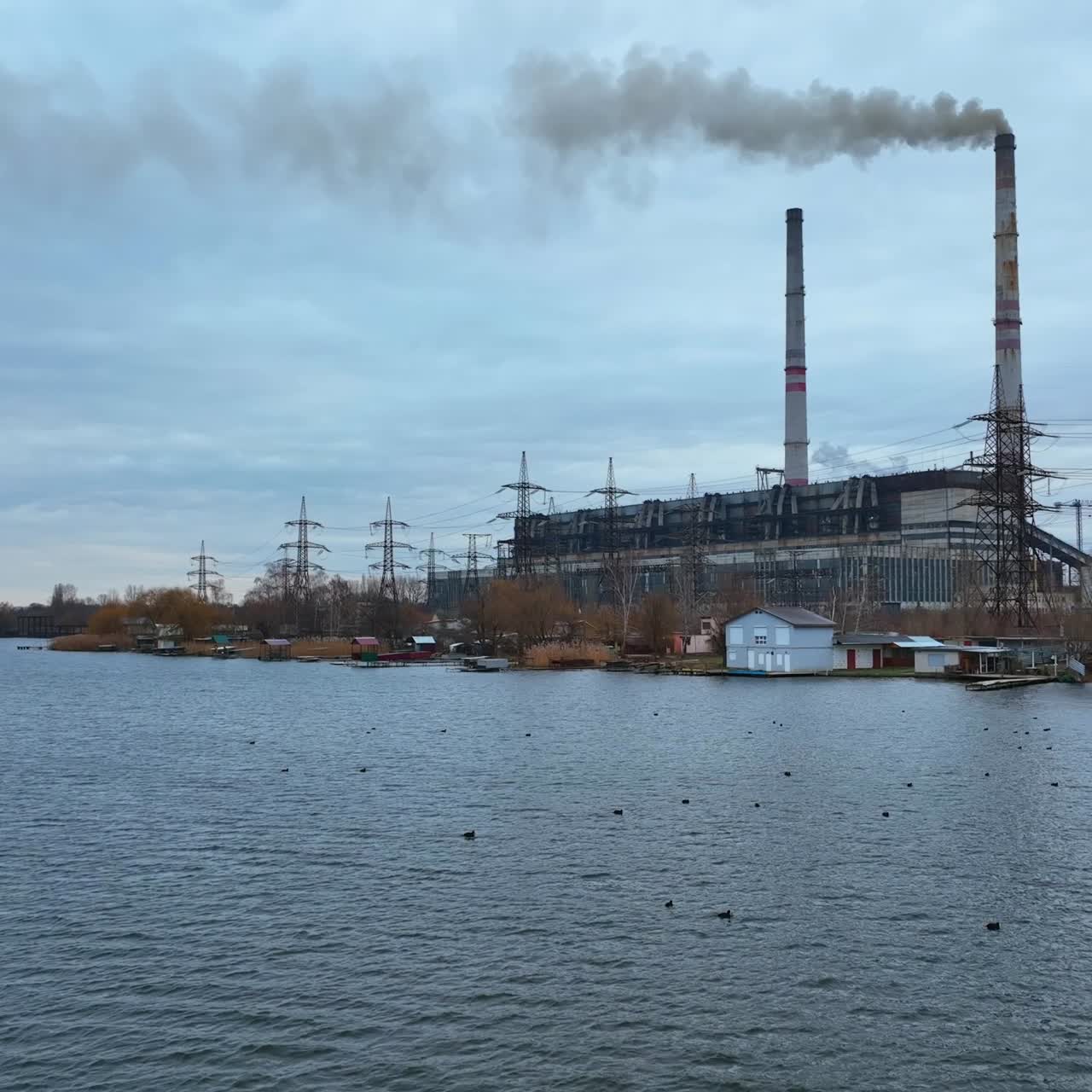 Industrial plant with two big flues rising in the air on the river bank. Dark smoke coming from the chimneys. Ducks floating on the river at the foreground