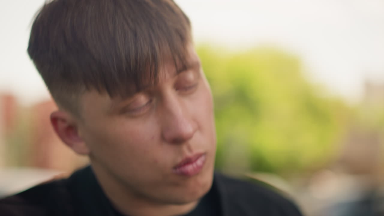 초록색 공원 밖에서 머리를 확인하는 백인 젊은 남성, 생각에 잠긴 몸짓, 앞머리를 빗어 넘기는 손가락, 불안하면서도 집중된 표정, 여름 햇살과 중요한 순간을 위한 조용한 준비