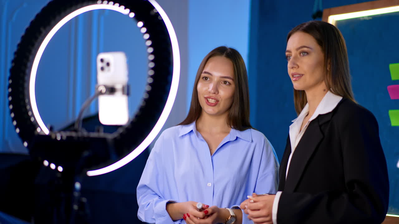 Attractive Caucasian brunettes stand at the ring light and smartphone. Bloggers talk and gesture to camera taking video content.