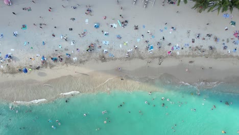 a drone hovers above a crowded tropical beach vacation destination as families enjoy sun, sand and surf together