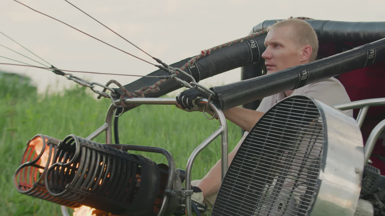 Man wearing gloves operating burner system as fire streams into balloon envelope, surrounded by cables and mechanical framework during hot air balloon setup in open grassy field