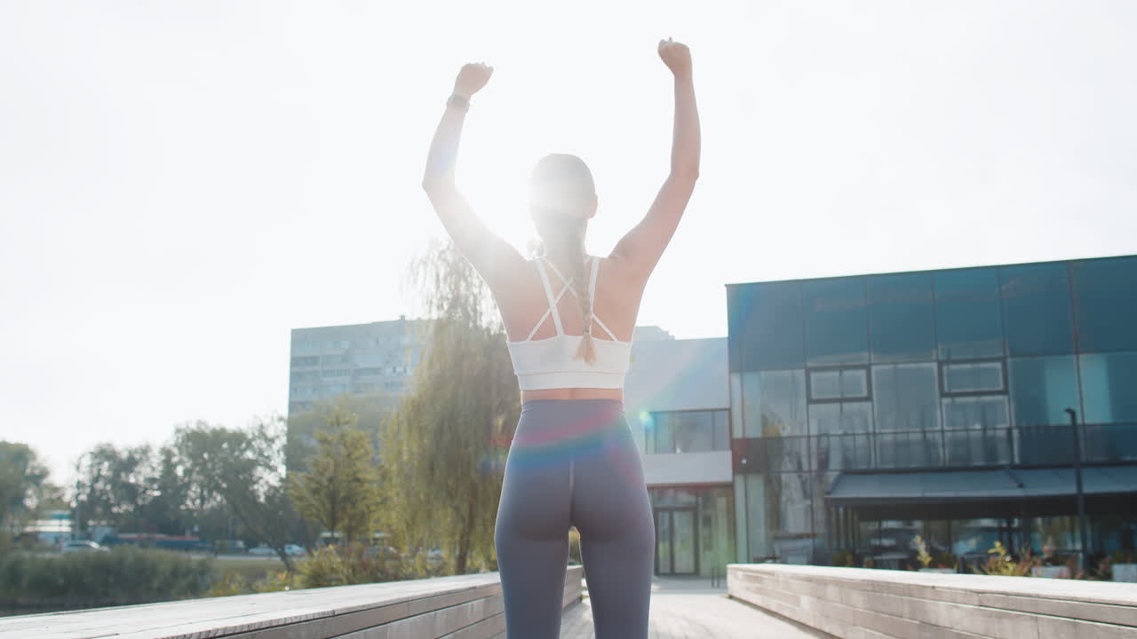 Rear view of young woman runner raising arms celebrating success after jogging in urban city park