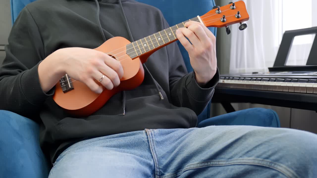 Man gently strumming ukulele strings indoors with shallow depth of field on hands, seated next to piano