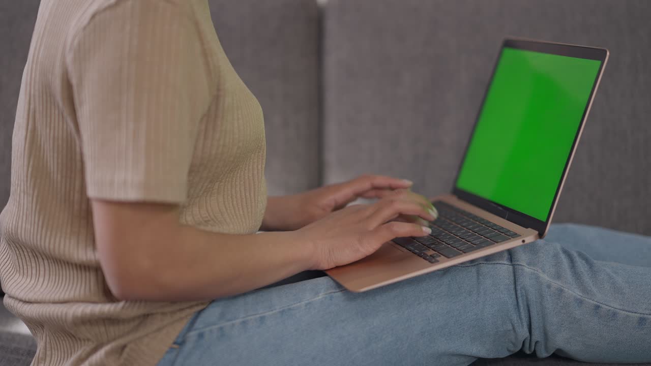 vista lateral de una mujer afroamericana irreconocible escribiendo en el teclado de la computadora portátil con pantalla verde. dispositivo chromakey en las piernas de una joven mujer delgada mensajería en línea navegación por internet en la oficina de casa.