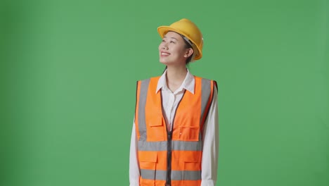 안전 헬을 입은 아시아 여성 엔지니어가 녹색 스크린 배경 스튜디오에 서서 주변을 둘러보고 있습니다.